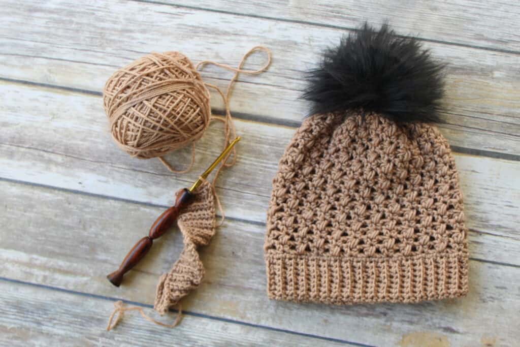 Photo of a taupe colored crochet hat, called the Appleton Hat, with a cake of yarn and crochet hook next to it. 