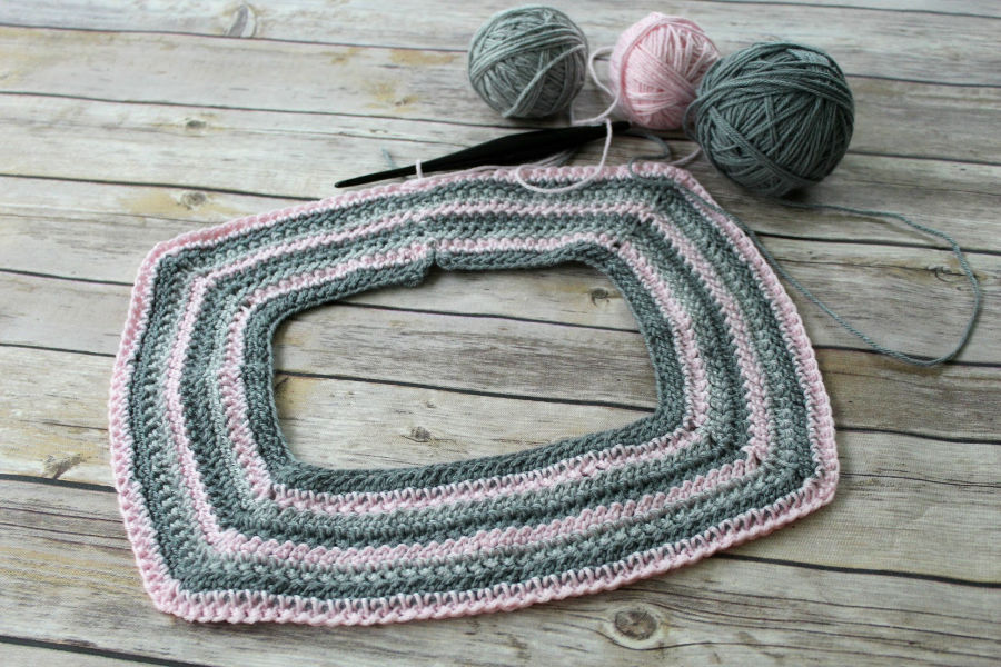 The start of a raglan crochet sweater using two shades of gray yarn and one shade of pink yarn, as well as a black crochet hook. 