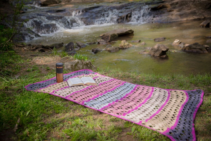 Serenity Blanket spread out on the grace with a bible and a travel mug on it in front of a stream. 