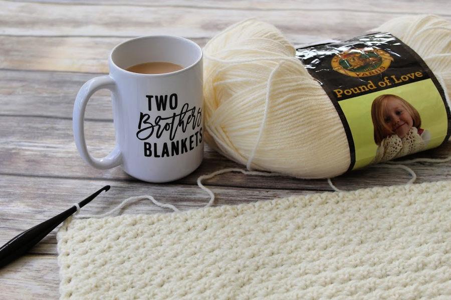 Crochet Lap Blanket being crocheted with skein of cream colored yarn and cup of coffee sitting next to it. 