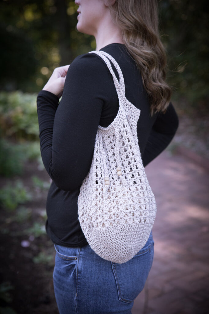 Woman carrying a tan colored crochet tote bag over her shoulder. This bag is called the Katie Tote. 
