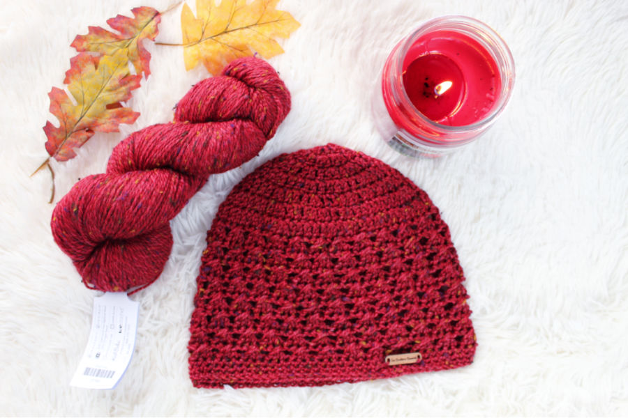 Flat lay photo of a red crochet beanie, called the Bonnie Bell Beanie. It is surrounded by a skein of red yarn, a red candle, and yellow and red fall leaves. 