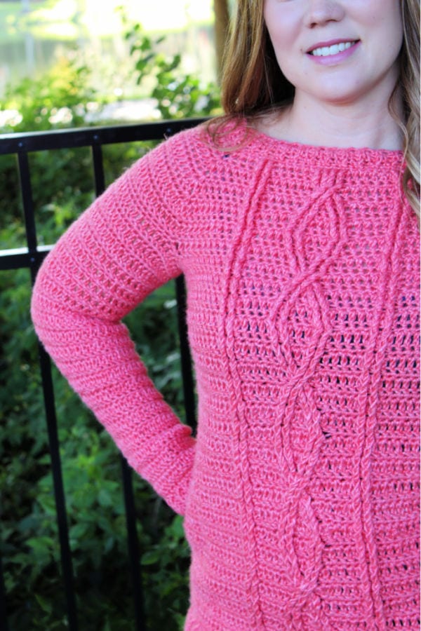 Close up of woman wearing a pink crochet sweater with cables. This sweater is called the Crossroads Sweater. 