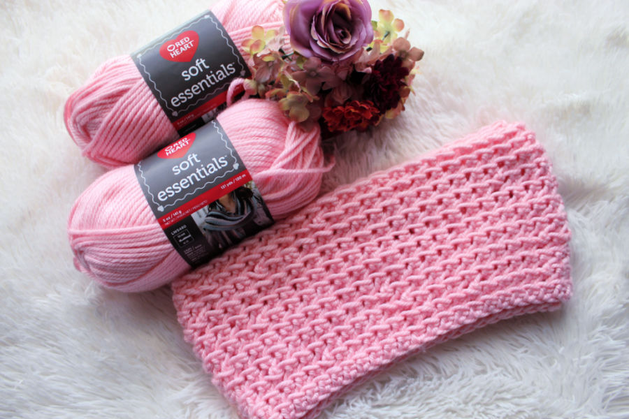 Flat lay photo of a pink crochet infinity scarf, called the KJ Infinity Scarf. The yarn used to make the scarf and a small bouquet of flowers is set beside the scarf. 