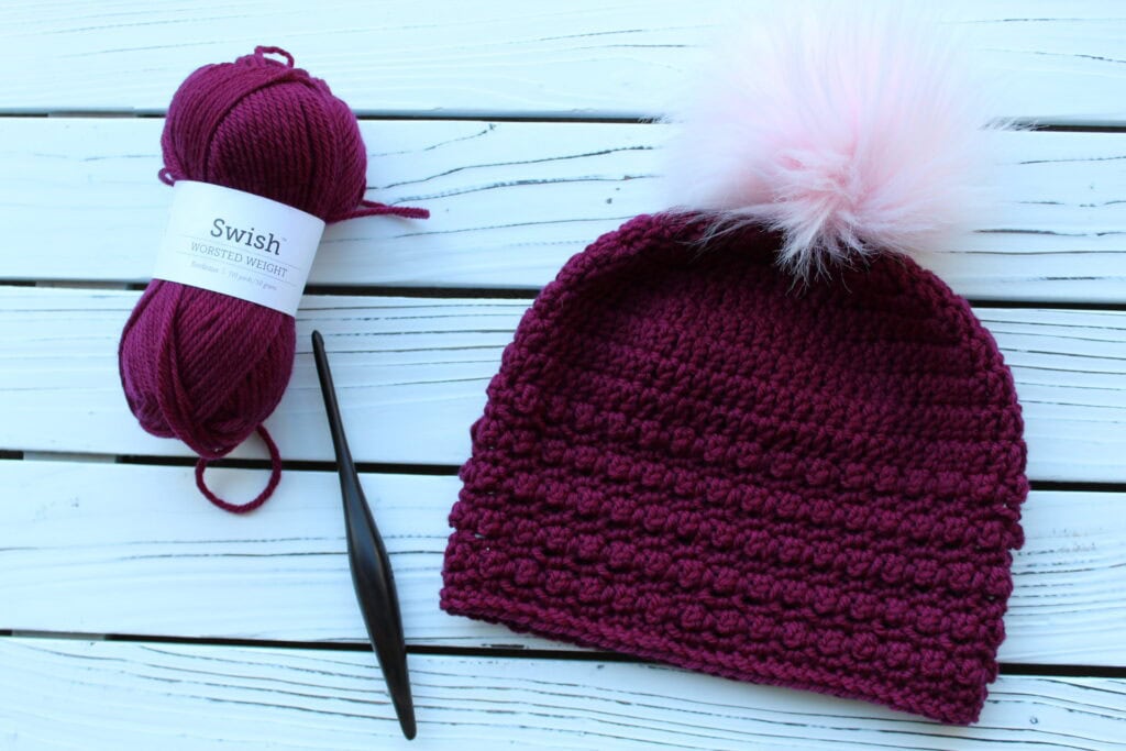 Flat lay of yarn, crochet hook, and crochet beanie in a dark pink color with a light pink pom. 