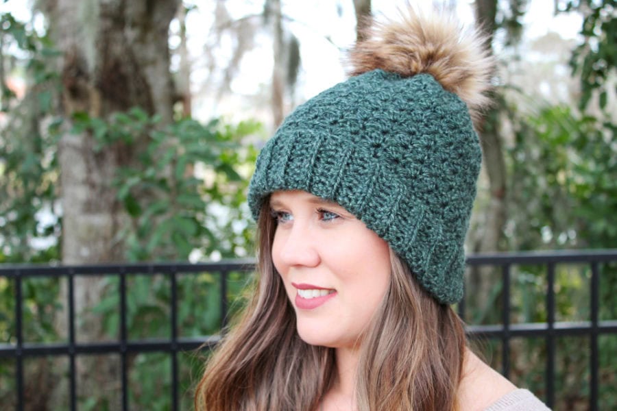 Woman wearing a green crochet hat with a brown pom. This hat is called the Ferguson Hat. 