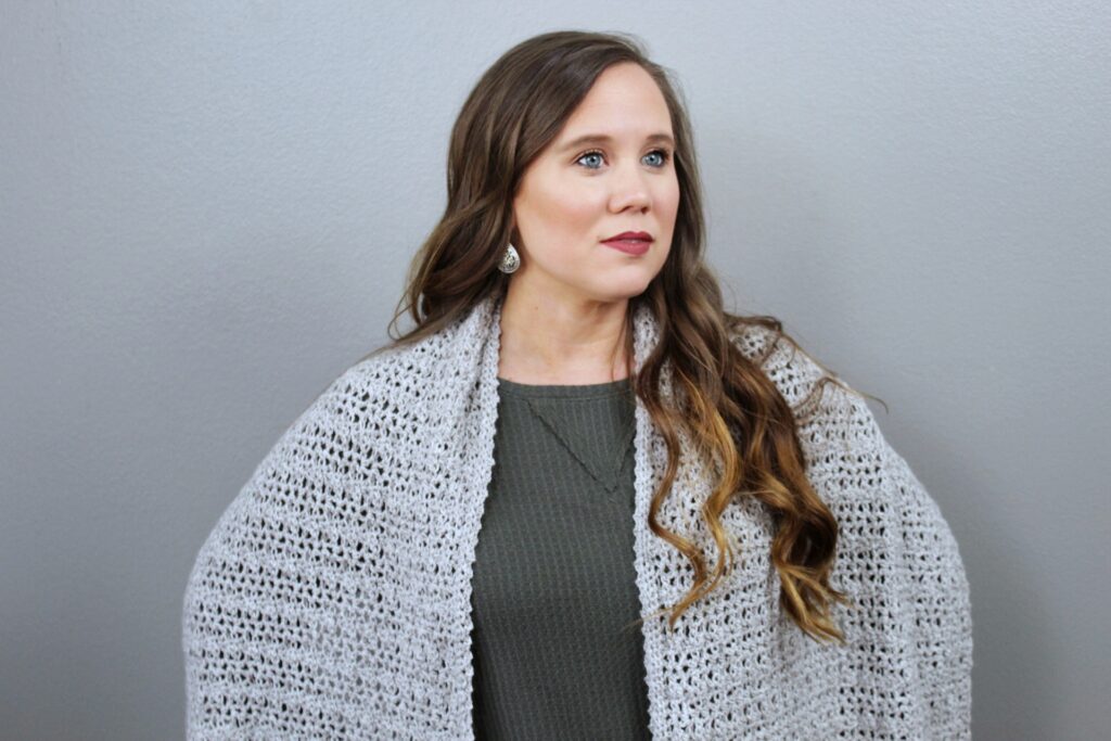 Woman wearing a long, light gray crochet wrap, called the Katie Wrap. 
