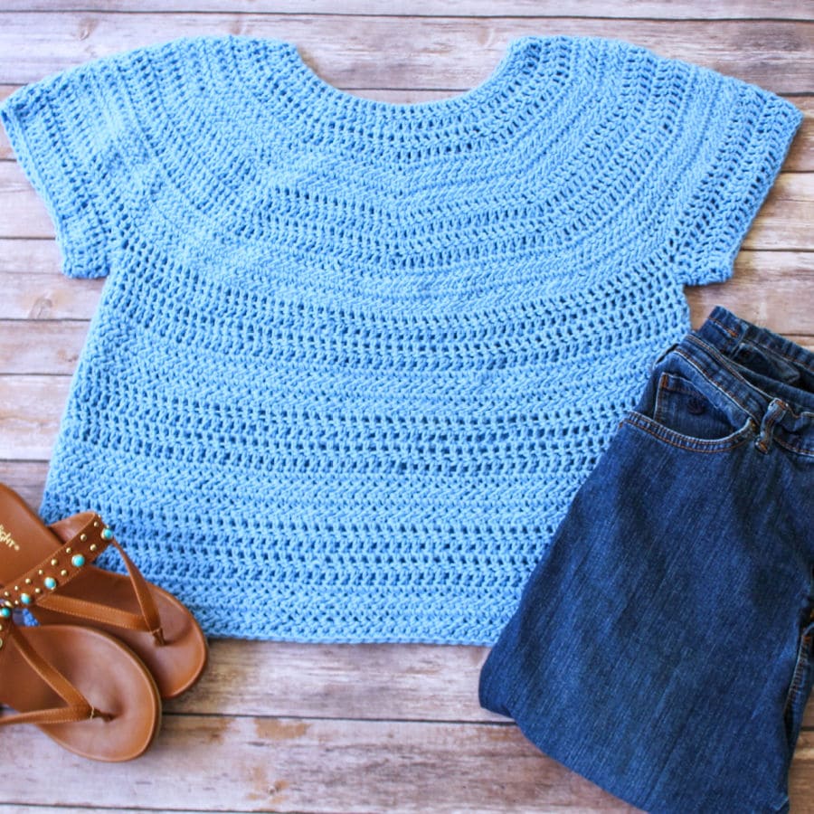 Flatlay photo of a blue short sleeved crochet top, called the Larkin Tee. Top is photographed with jeans and sandals. 