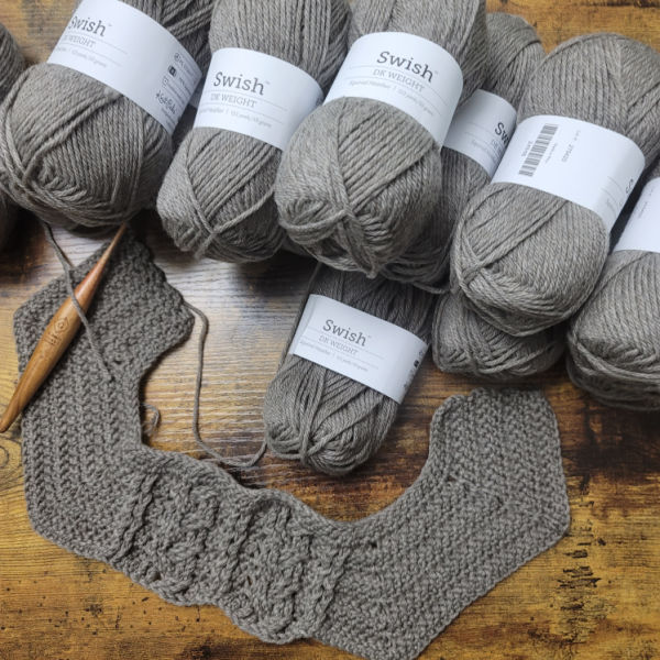 A photo of many skeins of gray Swish DK yarn, along with a yoke made with this yarn and a wooden crochet hook. 