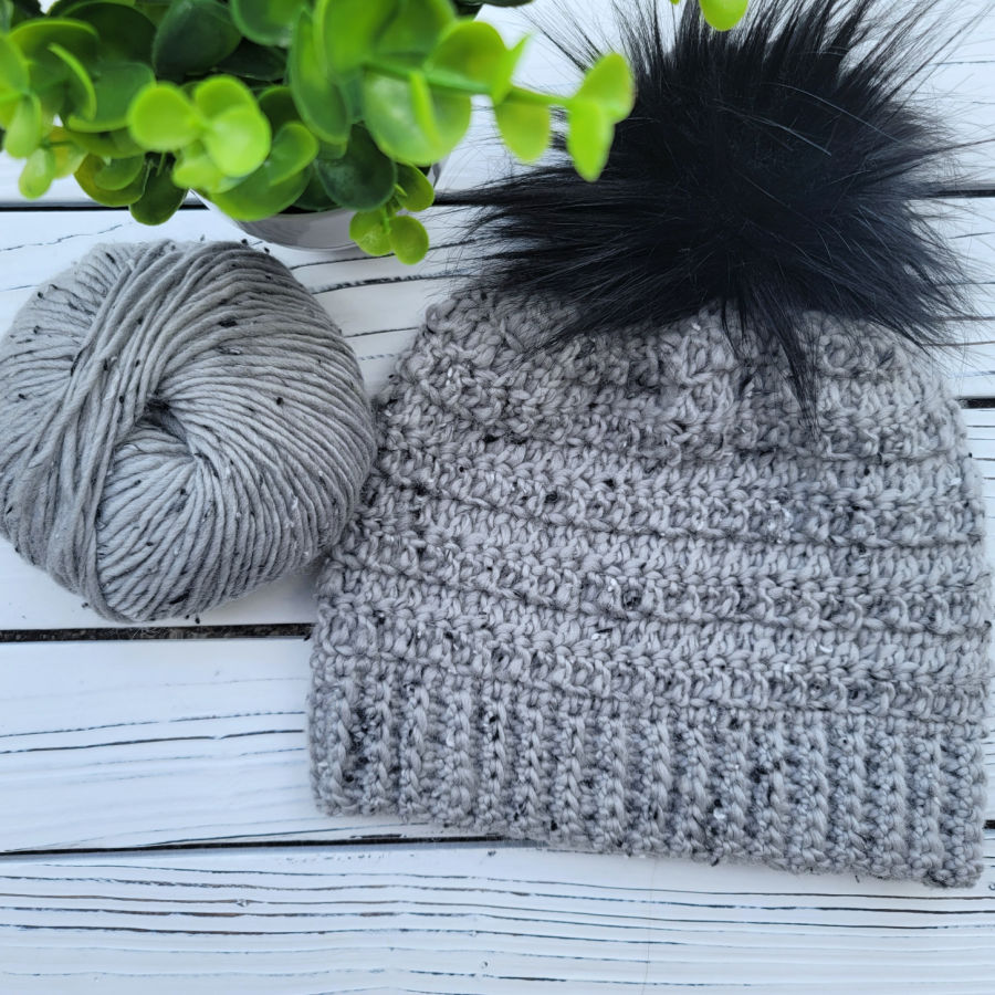 Flat lay image of a gray tweed crochet hat with a black pom attached. 