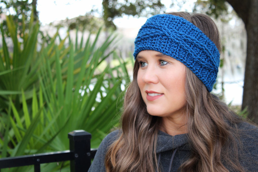 Woman wearing a blue cabled crochet headband, called the Crossroads Headband. 