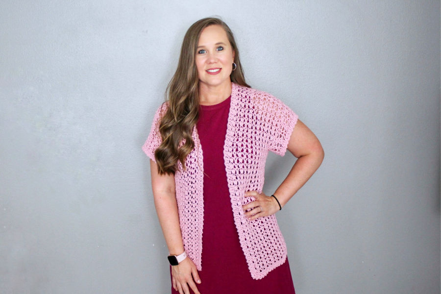 Woman wearing a pink, short sleeved crochet cardigan, called the Katie Cardi. 