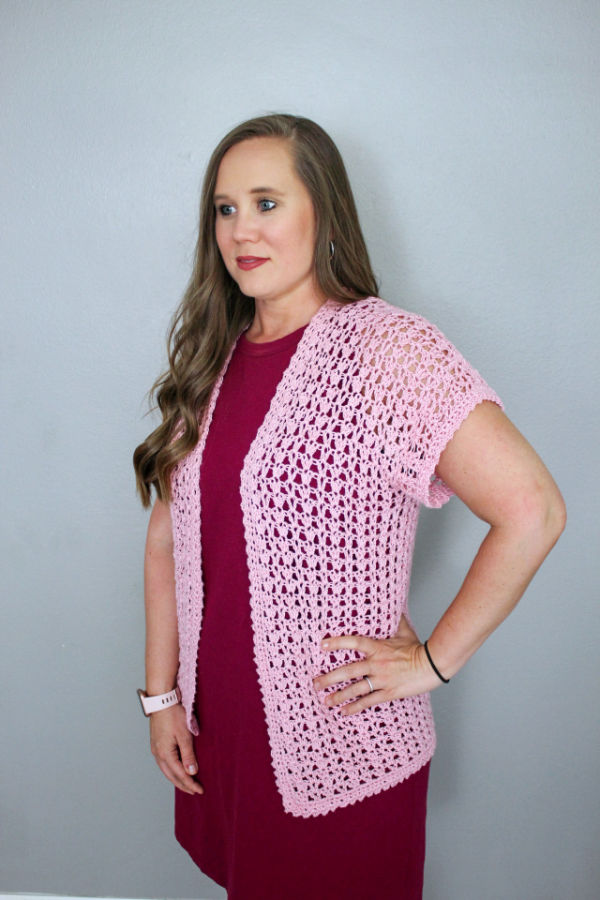 Woman wearing a pink, short sleeved crochet cardigan, called the Katie Cardi. 