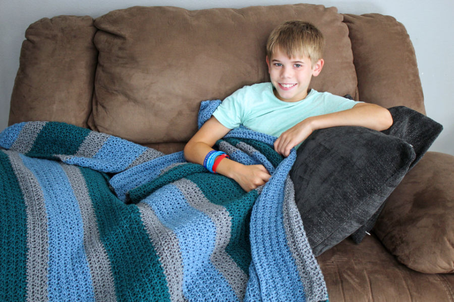 Boy with a large crochet blanket draped over him on the couch. 